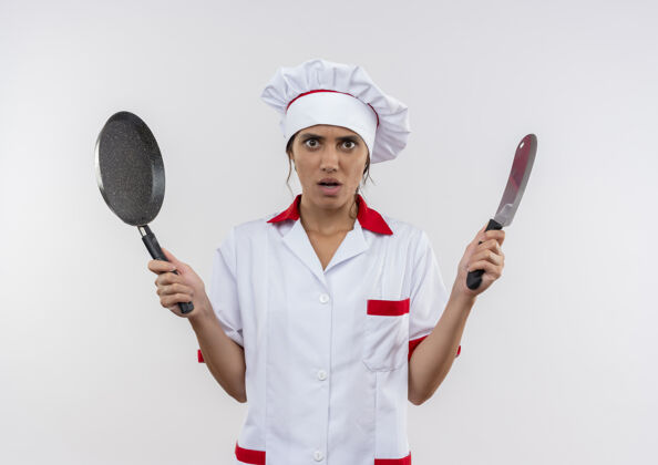 年轻年轻的女厨师穿着厨师制服 拿着菜刀和煎锅 站在隔离的白色墙壁上 留着复印空间平底锅拿着厨师