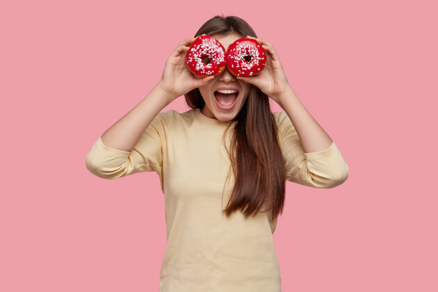 饥饿摄影棚拍摄的快乐的黑发女人用两个红色的甜甜圈遮住眼睛 精神抖擞 穿着黄色的衣服满意釉面年轻人