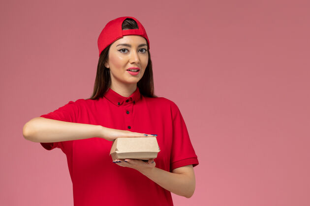 服务正面图：身穿红色制服和斗篷的年轻女快递员 手上拿着一个小小的快递食品包 放在粉红色的墙上送货女人前面