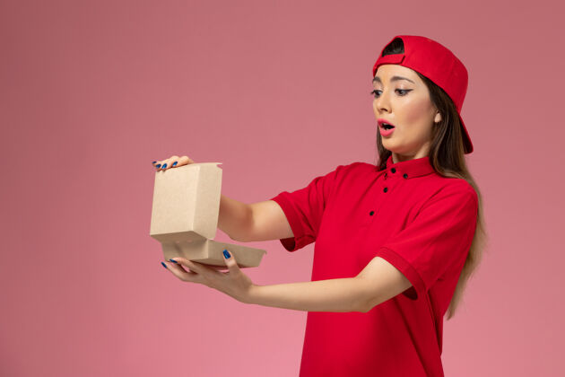 送货正面图：身穿红色制服和斗篷的年轻女快递员 手上拿着一个小小的快递食品包 放在淡粉色的墙上人抱着年轻