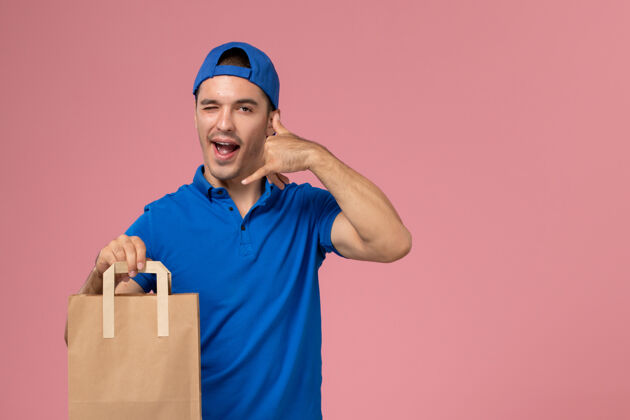 人正面图：身穿蓝色制服和披风的年轻男性快递员 手上拿着一个纸包裹 放在粉红色的墙上年轻送货人