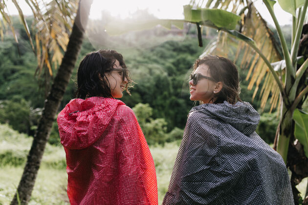 棕榈树有趣的女人湿头发看着对方 而走在森林里户外照片的女游客穿着雨衣站在大自然上户外放松热带