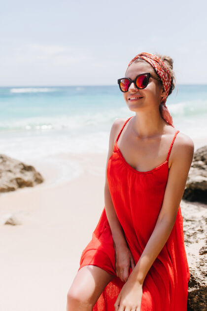 水坐在海滩上的笑容可掬的晒黑的白人女子穿着红色连衣裙在海边休闲的白种人女模特的户外写真海洋风景时尚